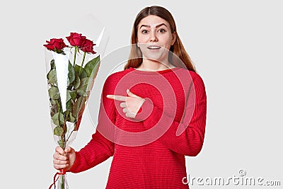 Surprised beautiful woman with cheerful expression, dark eyebrows, points at beautiful bouquet of roses, wonders recieving present Stock Photo