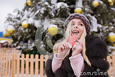 Surprised young woman dressed in fur coat and knitted hat, eating tasty christmas gingerbread against light decoration Stock Photo