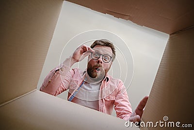 Surprised man looks into the unpacked delivered box with the order, hold glasses with hands and rejoices. Unboxing Stock Photo