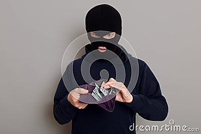 Surprised man thief dressed in robber mask holding open wallet in his hands, shocked by what he sees, steals big sum of money, Stock Photo