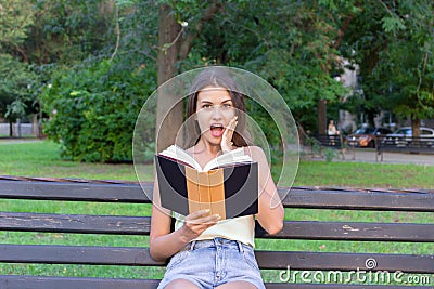 Surprised young woman with widely open yeas and mouth and a hand on cheek is reading a book outdoors Stock Photo