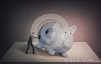 Surreal business concept, tiny businessman trying to push a huge piggybank out of the table, to get something out of it. Breaking Stock Photo