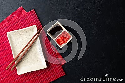 Sushi chopsticks, soy sauce, ginger, red bamboo mat and empty plate on black background. Stock Photo