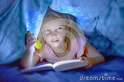 Sweet beautiful and pretty little blond child girl 6 to 8 years old lying under bed covers reading book in the dark with torch fla Stock Photo