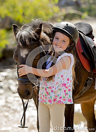 Sweet beautiful young girl 7 or 8 years old hugging head of little pony horse smiling happy wearing safety jockey helmet in summer Stock Photo