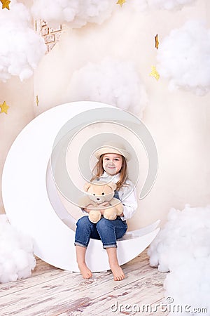 Sweet Dreams. Little cute girl sitting on the moon with clouds and stars with a teddy bear in their hands and playing. Little astr Stock Photo