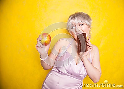 Sweet joyous curvaceous girl with a short haircut stands in a delicate evening silk dress and compares bad and good food Stock Photo