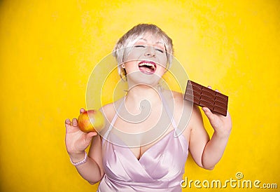 Sweet joyous curvaceous girl with a short haircut stands in a delicate evening silk dress and compares bad and good food Stock Photo