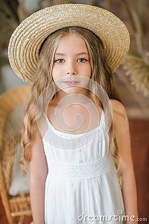 A sweet little girl with long blond hair in a white sarafan and a straw hat in a rattan chair Stock Photo