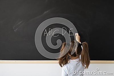 Sweet little girl at school in a lesson. A child stands in front of a black board. She writes in English the letters of Stock Photo