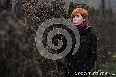 Sweet red-haired girl in a black coat and purple knitted scarf is standing by the fence overgrown with grapevine or ivy Stock Photo