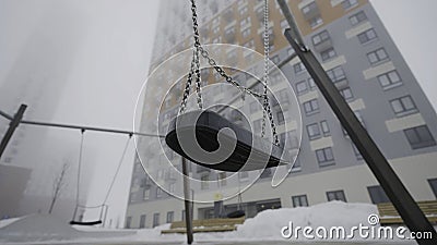 Swing on background of houses in fog. Action. Empty swing eerily seesaw on background of fog. Empty playground in cloudy Stock Photo