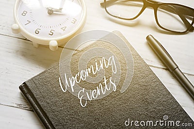 Table clock,eyeglasses,pen and notebook written with UPCOMING EVENTS on white wooden background with sun flare Stock Photo