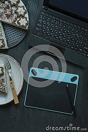 On the table is a laptop, graphics tablet, cake and a cup of coffee. Office supplies. Working environment. View from above. Dark Stock Photo