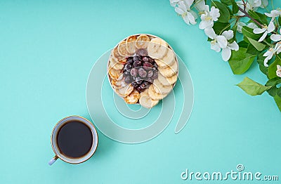 On the table is a plate with mini-pancakes, bananas and frozen currants, a cup of coffee and a branch of a blossoming apple tree. Stock Photo