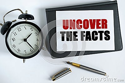On the table there is a clock, a pen, a notebook and a card on which the text is written - UNCOVER THE FACTS Stock Photo