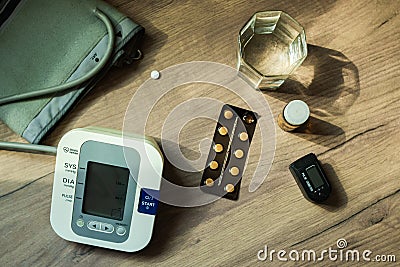 On the table there are pills, a blood pressure measuring device, a glass of water and an oxyometer. An elderly person`s kit. Top Stock Photo