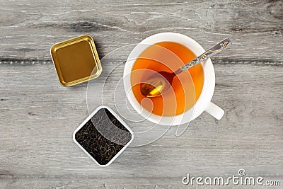 Table top view on cup of hot amber tea with spoon, and caddy full of black dried loose leaves, placed on gray wood desk. Stock Photo