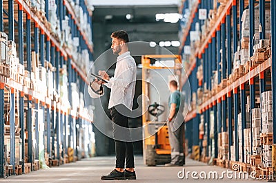 With tablet in hands. Storage worker is in the warehouse with bunch of products Stock Photo