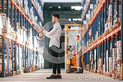 With tablet in hands. Storage worker is in the warehouse with bunch of products Stock Photo