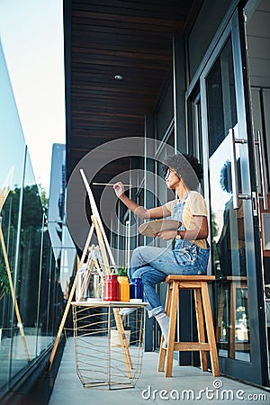Take some time to sit down and get artsy. a young artist painting on a canvas while sitting on the balcony at home. Stock Photo