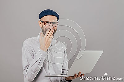 Talented male graphic designer works remotely on gadget, laughs positively, covers mouth with palm, wears spectacles and white Stock Photo