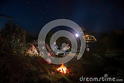 They talk and drink. car and hikers near campfire. Couple man and woman sitting near bonfire under majestic blue sky with stars. Stock Photo