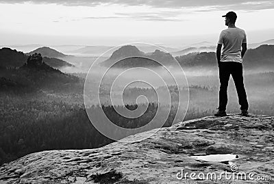 Tall man in white shirt and black trousers with red baseball cap stay on sharp cliff and watch to valley. Colorful misty mornin Stock Photo