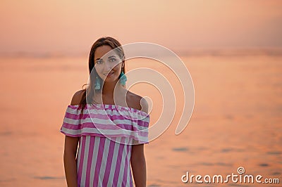 Tanned beautiful woman in dress with white and pink stripes. Close up and copy space. Orange sea or ocean at the sunset. Vacations Stock Photo