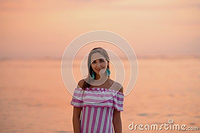 Tanned beautiful woman in dress with white and pink stripes. Orange sea or ocean at the sunset. Vacations concept Stock Photo