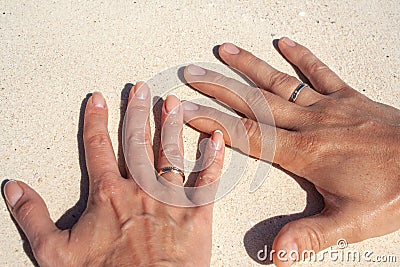 Tanned hands of newlyweds with gold rings on ring fingers on white sand as a symbol of wedding travel Stock Photo
