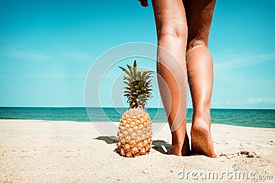 Tanned legs of young woman standing with pineapple at tropical beach in summer Stock Photo