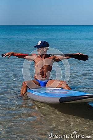 Tanned sporty man in a cap sits on his surfboard on the water holding by hands an oar behind his head and looks into the Stock Photo