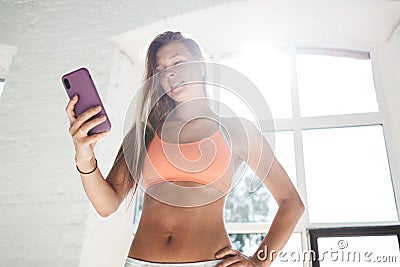 Tanned sporty woman wearing sportwear and wireless headphones in white sunny gym hold smartphone in hand and counts calories in fi Stock Photo