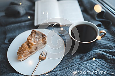 Tasty apple pie on white plate with cup of black tea and open paper book on knit woolen cloth fabric closeup over Christmas glow l Stock Photo