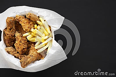 Tasty fastfood: fried chicken drumsticks, spicy wings, French fries and chicken fingers in paper box over black background, top vi Stock Photo