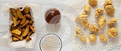 Tasty fastfood: fried potato wedges in paper box, chicken bites, barbecue sauce and beer on a white wooden table, top view. Flat Stock Photo