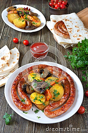 Tasty homemade grilled sausages with baked herb potatoes, mushrooms, cherry tomatoes and ketchup on a wooden background. Stock Photo