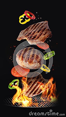 Tasty roasted meat and vegetables falling on barbecue grill with burning fire against background Stock Photo