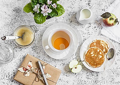 Tea, pancake, apple sauce, homemade gift in kraft paper, flower violet on a light background. Romantic breakfast table. Stock Photo