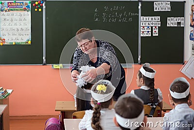 Teacher adjusts the bow on the schoolgirl, who sits at her desk, against the background of the school board in the school class. Editorial Stock Photo