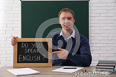 Teacher holding small chalkboard with inscription Do You Speak English? at table in classroom Stock Photo