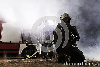 Team of professional firefighters holding water hose in front of a firetruck with smoke in the air. Half silhouette Stock Photo