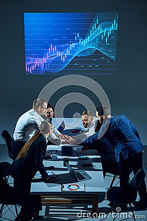 Team shakings hands for good cooperation. Office workers sit around conference table with digital stock chart displayed Stock Photo