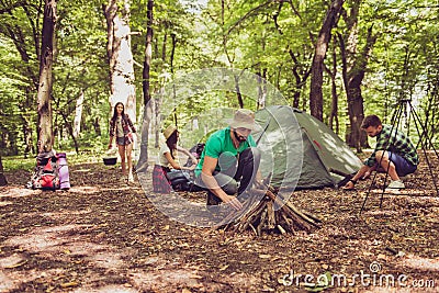 Team work. Young bearded male tourist in a hat is setting campfire, other set on a tent, ladys are unoacking and organizing the m Stock Photo