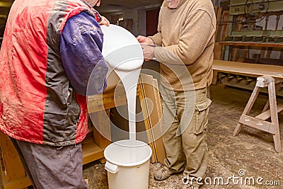 Two carpentry workers pour wood glue from a large plastic bucket into another empty Stock Photo