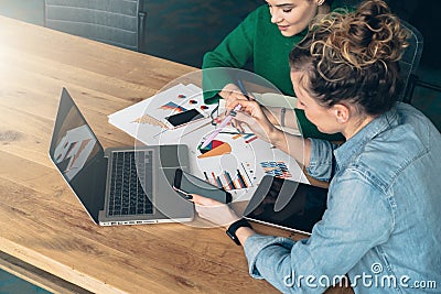 Teamwork. Two young business women sitting at table in front of laptop. On table is tablet computer and paper charts. Stock Photo