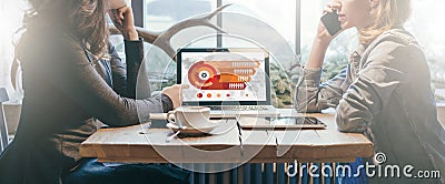 Teamwork, two young businesswomen sitting across table from each other. On table laptop, coffee cup and tablet computer. Stock Photo