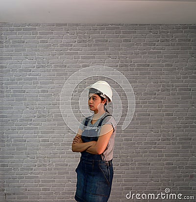 Technician woman ware white helmet with grey T-shirt and denim jeans apron dress standing forty five angle and hugging chest. Stock Photo