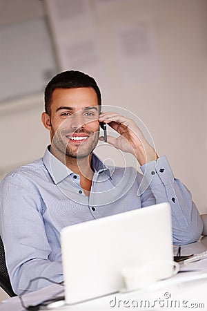 Technology keeps him at the top of his game. a smiling young man sitting at his desk and using his mobile phone. Stock Photo